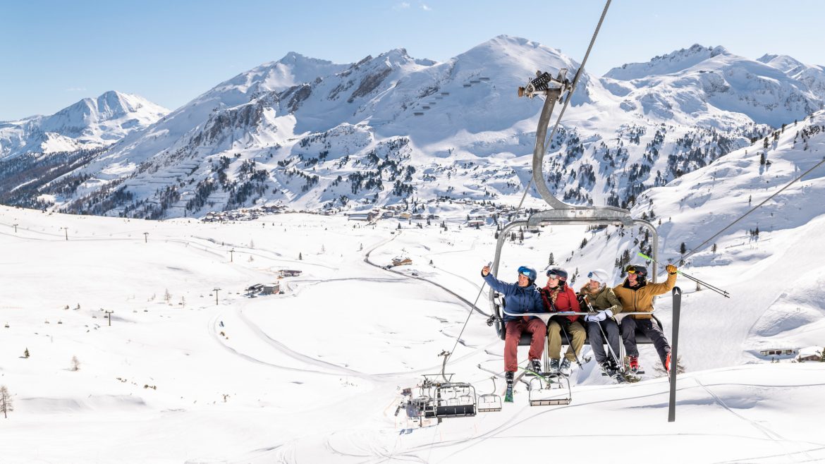 Vier Skifahrer fahren an einem hellen, klaren Tag mit einem Skilift über verschneite Berge.