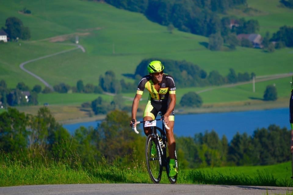 Radfahrer mit gelbem Helm fährt bergauf auf einer Landstraße mit grünen Hügeln und einem See im Hintergrund.
