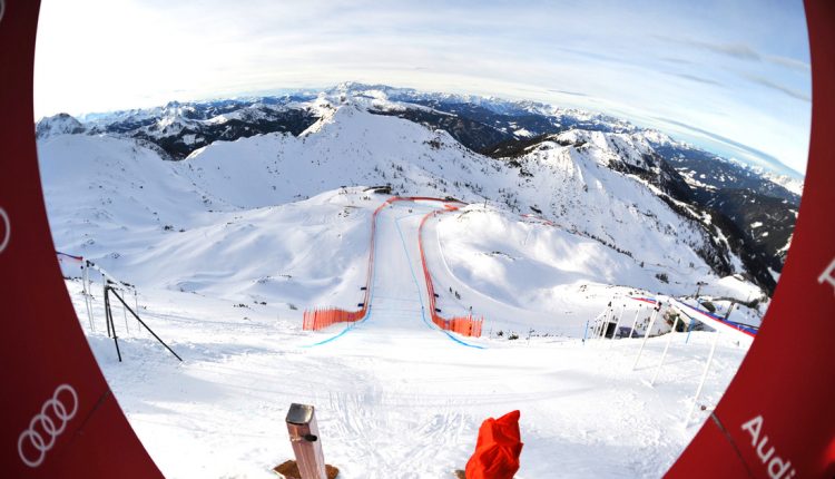 Blick durch einen runden Rahmen auf eine verschneite Skipiste, die sich einen Berg hinunterschlängelt, umgeben von schneebedeckten Gipfeln.