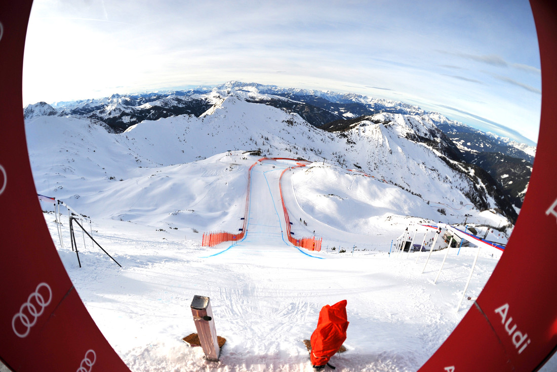 Blick durch einen runden Rahmen auf eine verschneite Skipiste, die sich einen Berg hinunterschlängelt, umgeben von schneebedeckten Gipfeln.