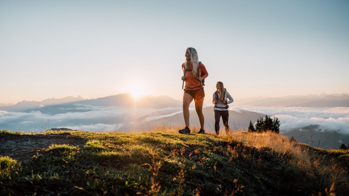 Zwei Frauen wandern auf einem grasbewachsenen Hügel bei Sonnenaufgang, mit Bergen und Wolken im Hintergrund.
