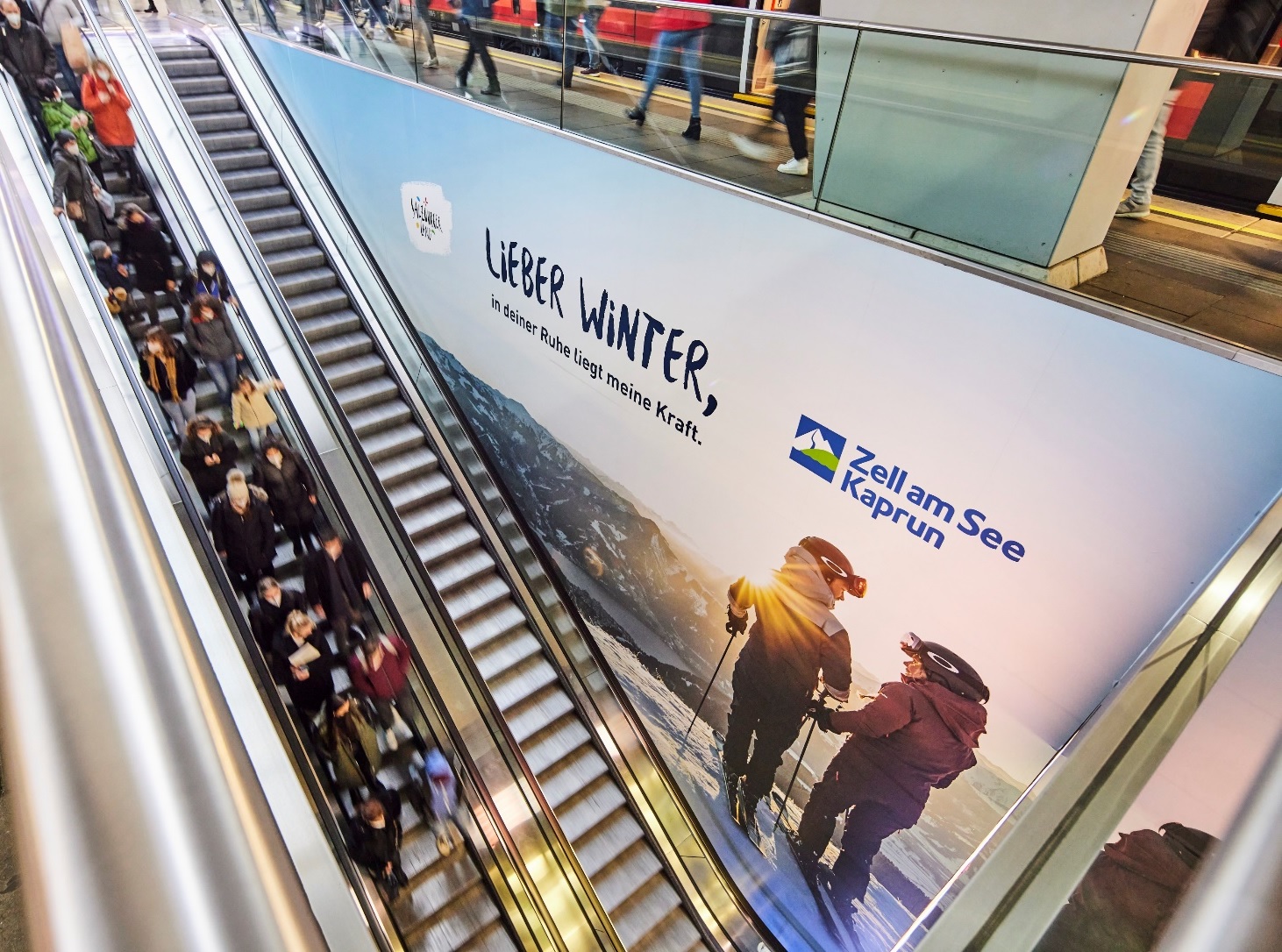 Menschen auf einer Rolltreppe neben einer großen Winterreisewerbung für Zell am See-Kaprun.
