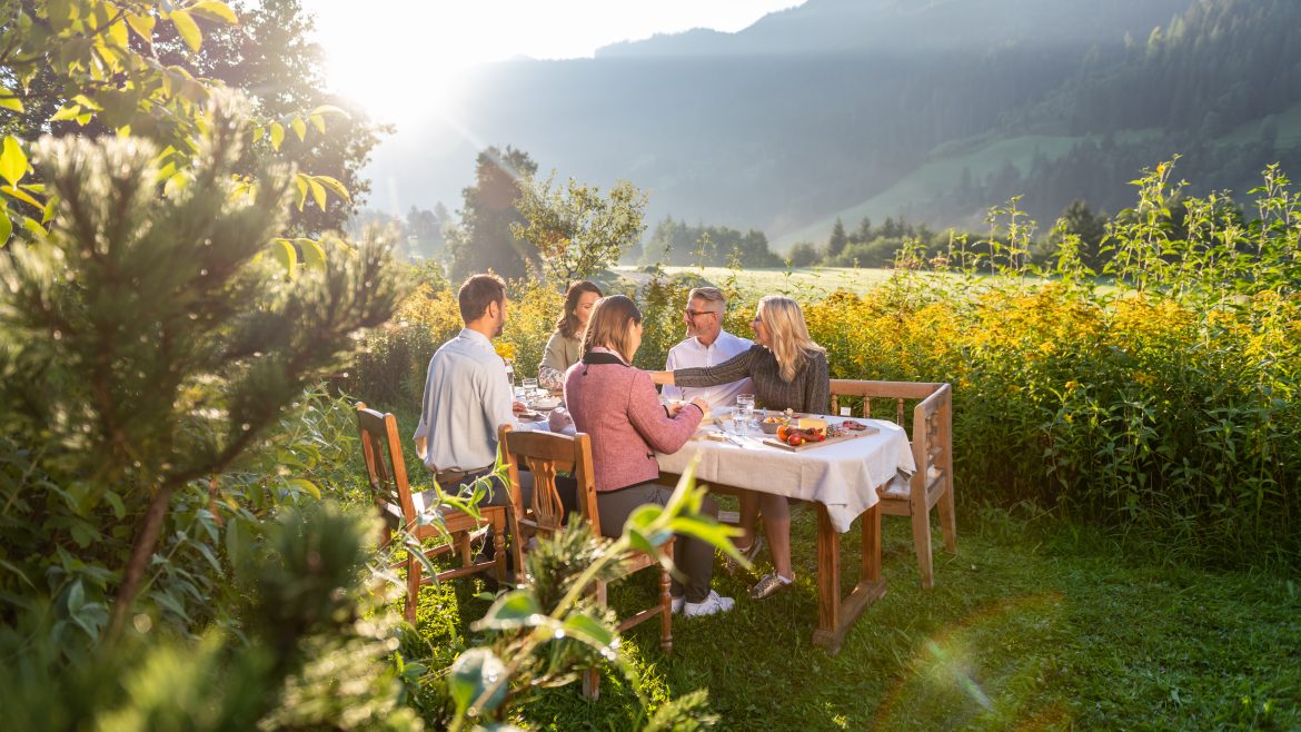 Fünf Personen speisen im Freien an einem Tisch in einer sonnigen, grünen Berglandschaft mit Bäumen und Hügeln im Hintergrund.