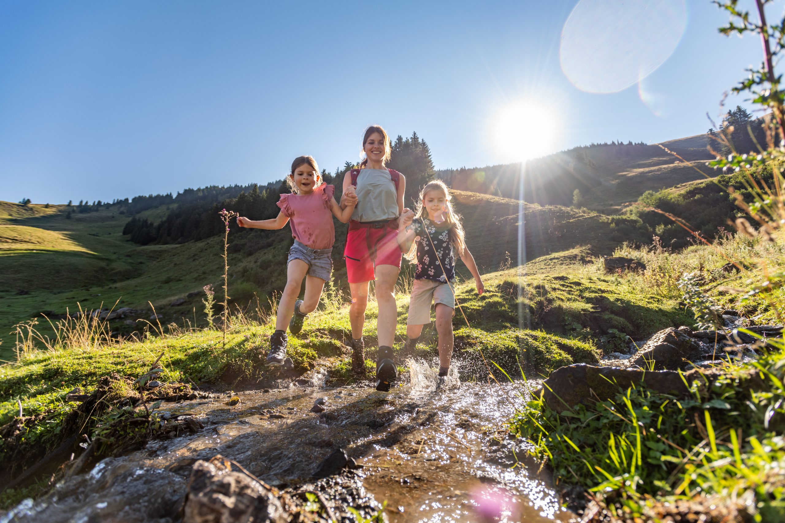 Eine Frau und zwei Mädchen wandern über einen kleinen Bach in einer sonnigen, grünen Berglandschaft.