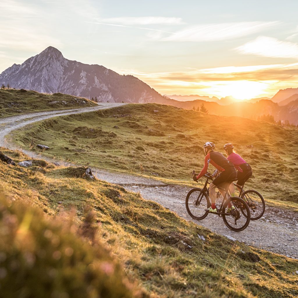 Zwei Radfahrer fahren bei Sonnenuntergang auf einem kurvenreichen Bergweg mit Bergen im Hintergrund.