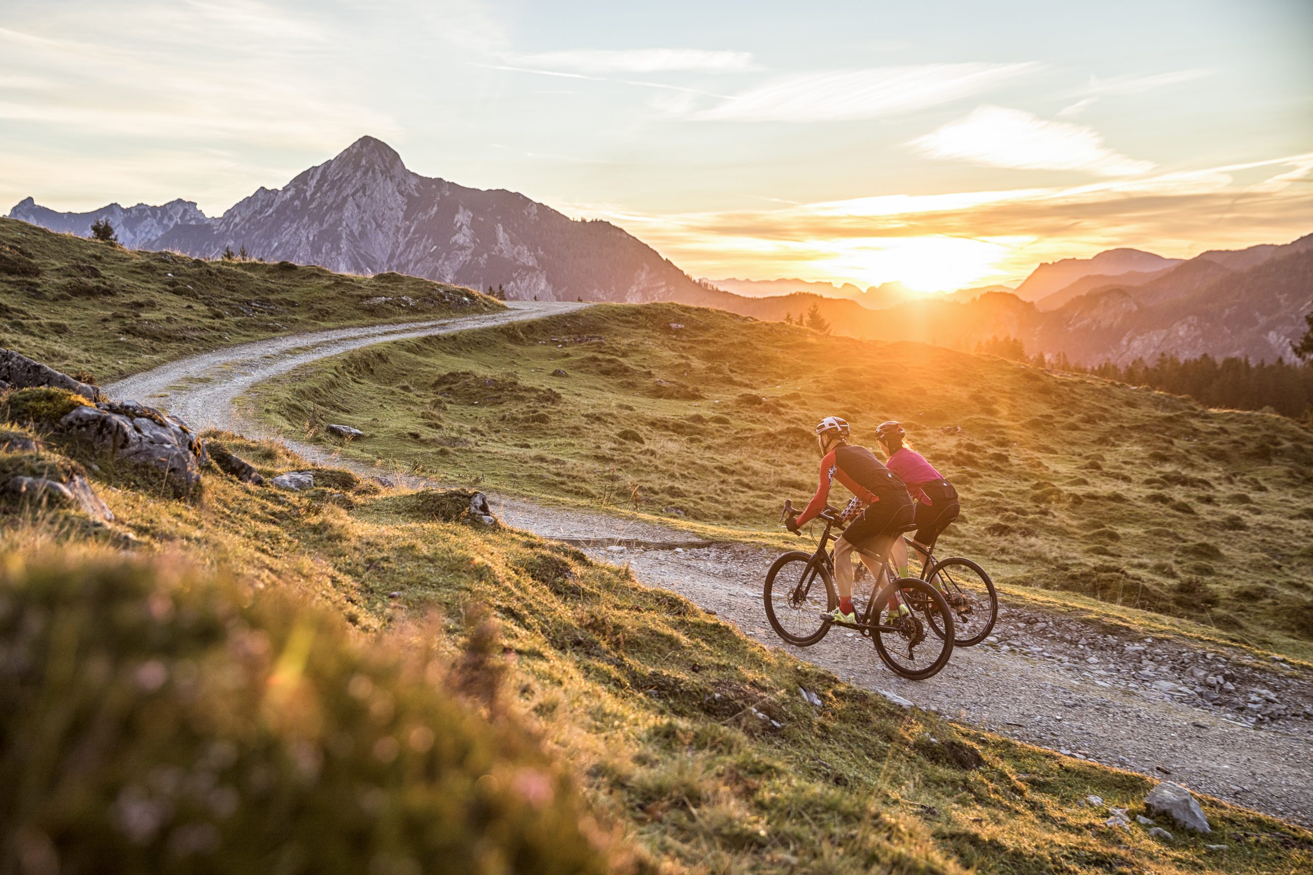 Zwei Radfahrer fahren bei Sonnenuntergang auf einem kurvenreichen Bergweg mit Bergen im Hintergrund.