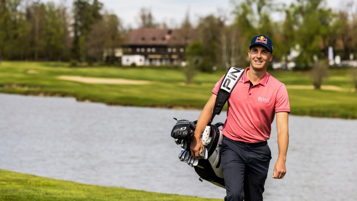 Ein lächelnder Golfer in einem rosa Hemd trägt eine Golftasche neben einem See auf einem üppigen, grünen Golfplatz.
