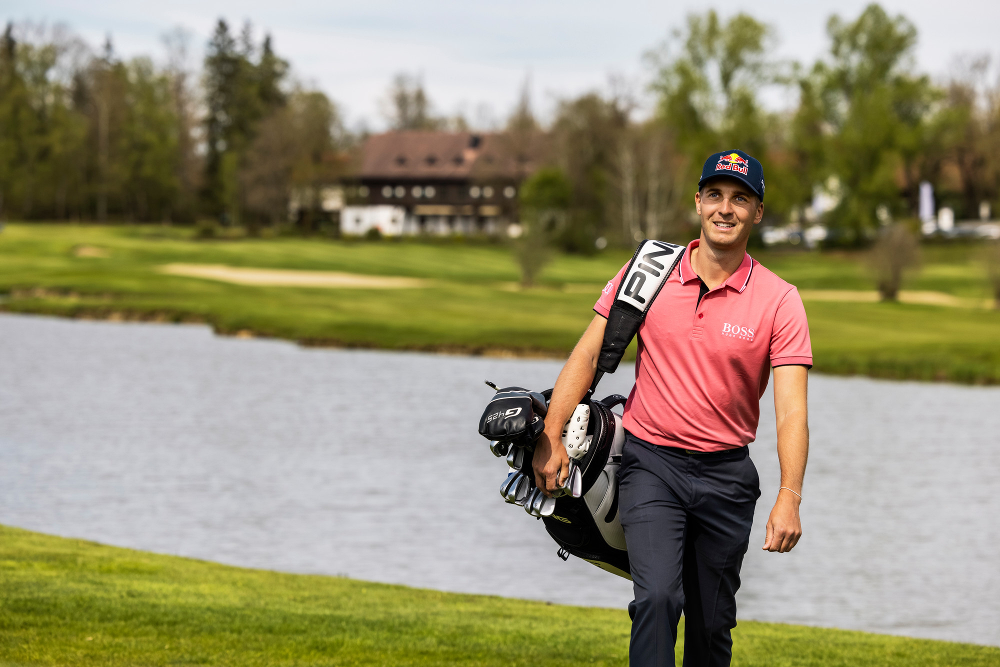 Ein lächelnder Golfer in einem rosa Hemd trägt eine Golftasche neben einem See auf einem üppigen, grünen Golfplatz.