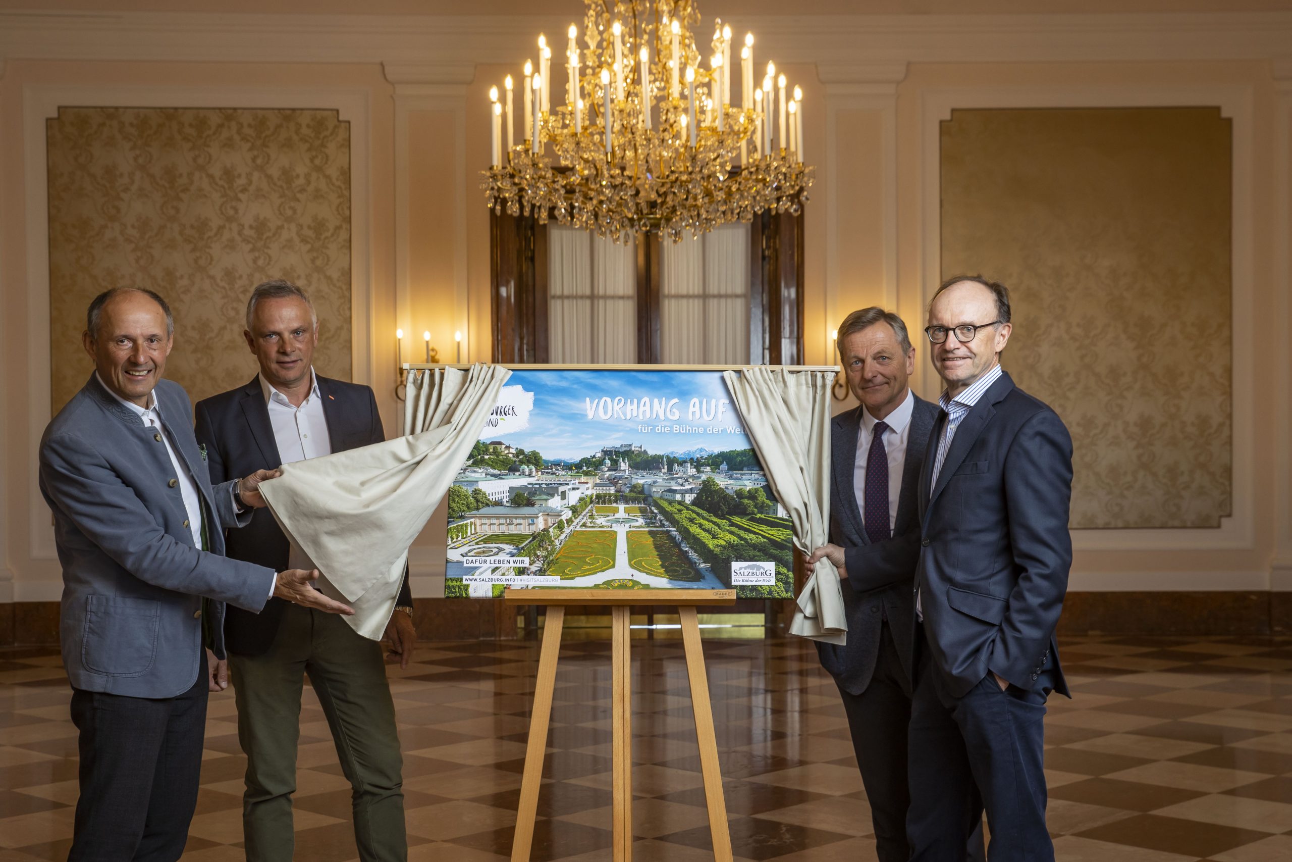 Vier Männer enthüllen in einem eleganten Raum mit Kronleuchtern und Marmorböden ein Poster mit Architekturplänen.