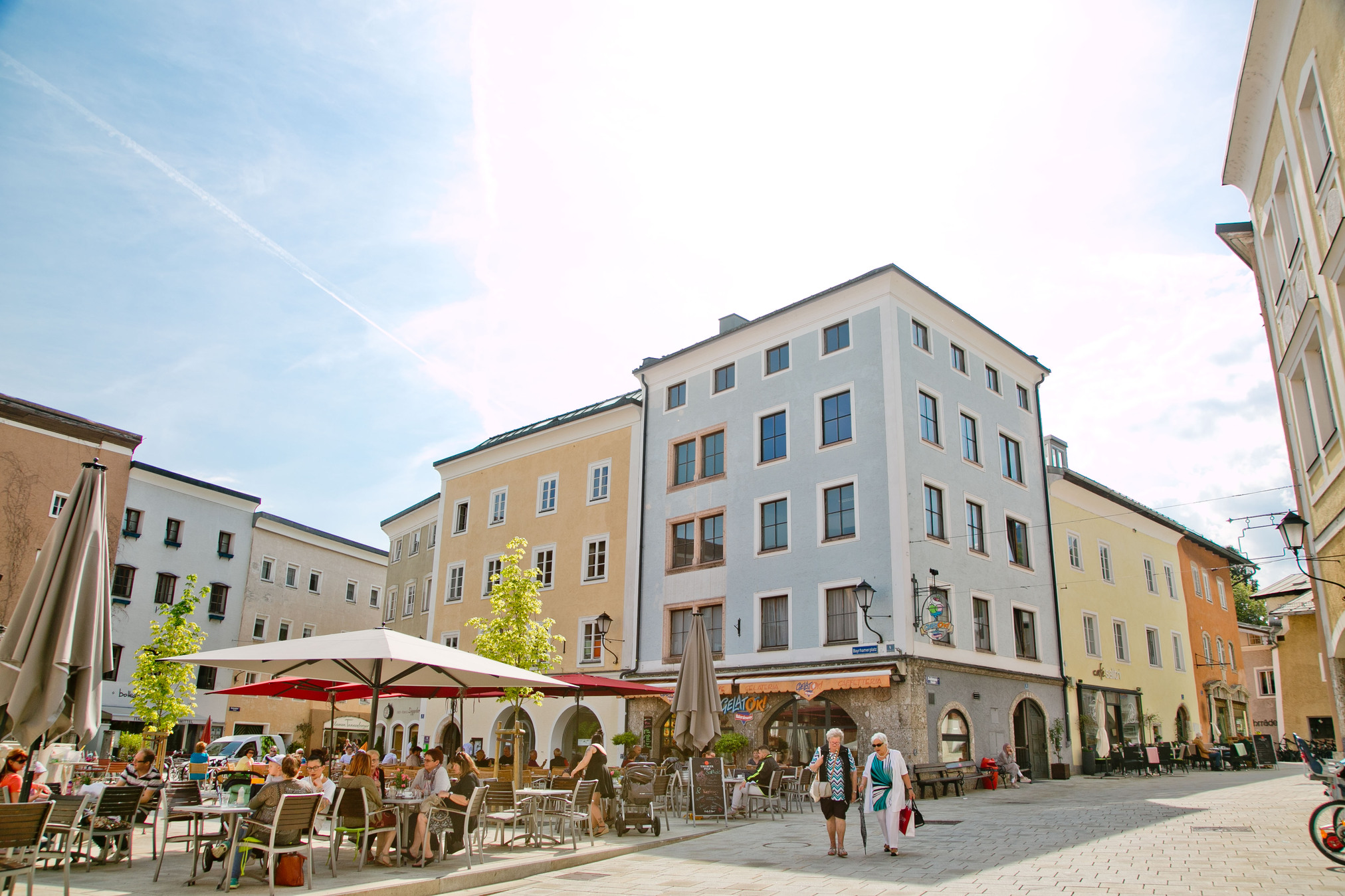 Ein Straßencafé auf einem europäischen Platz mit Menschen, die dort essen, Sonnenschirmen und pastellfarbenen Gebäuden an einem sonnigen Tag.