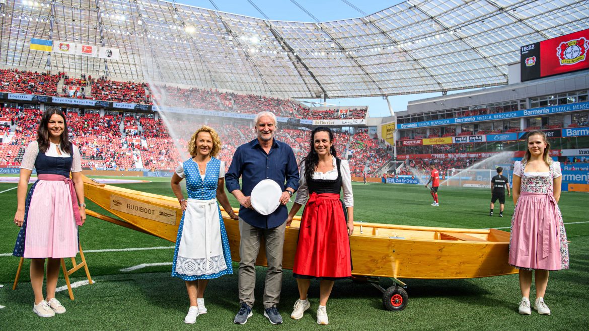 Fünf traditionell gekleidete Personen stehen auf einem Fußballfeld vor einem Holzboot mit Fans in Stadionsitzen.