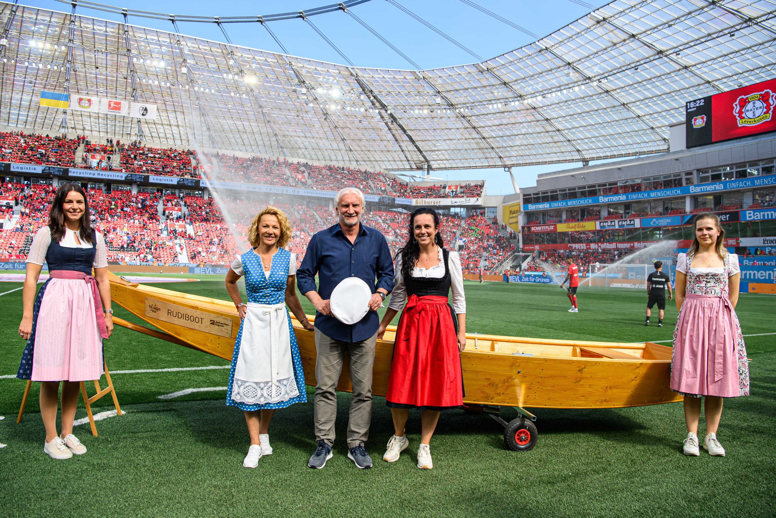 Fünf traditionell gekleidete Personen stehen auf einem Fußballfeld vor einem Holzboot mit Fans in Stadionsitzen.