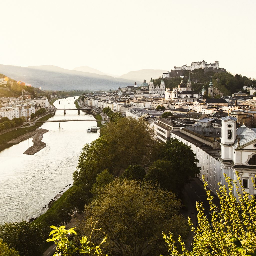 Eine malerische Ansicht von Salzburg, Österreich, mit einem Fluss, Brücken und historischen Gebäuden bei Sonnenuntergang.