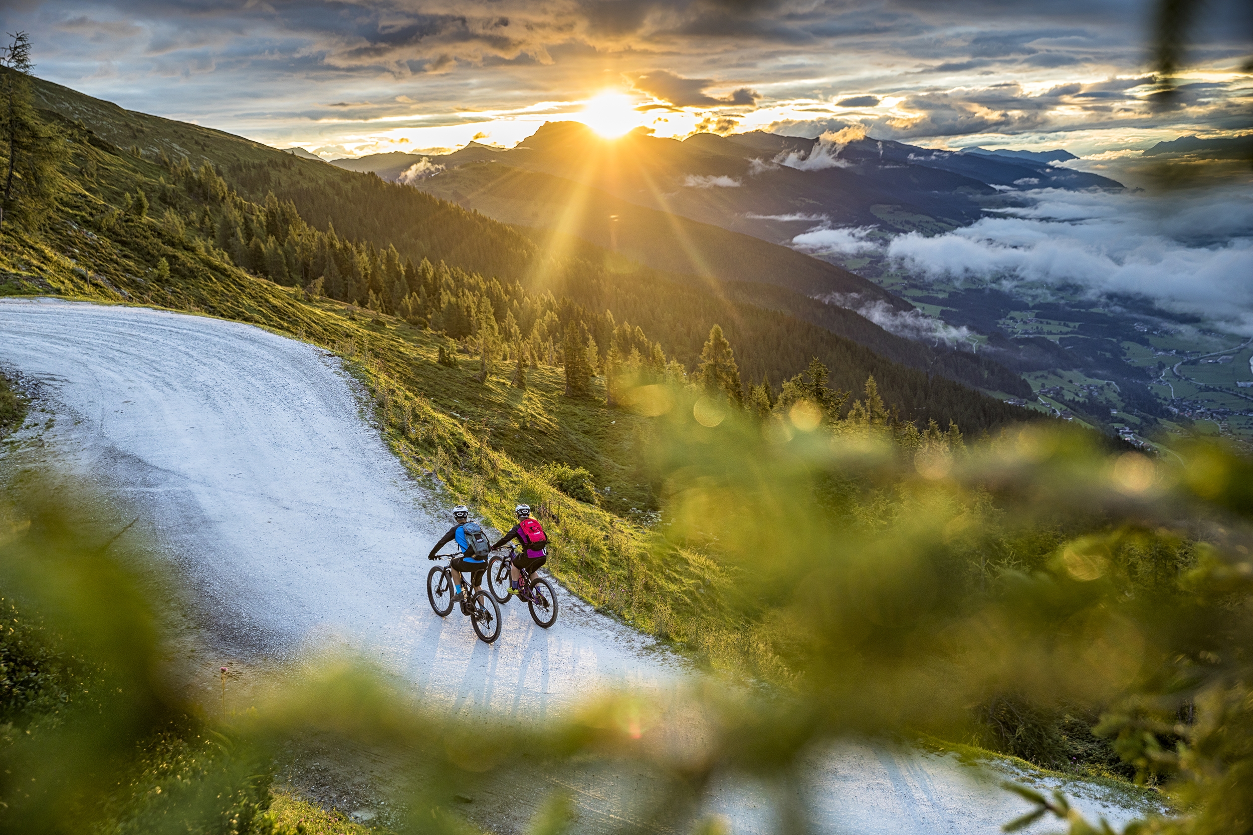 Zwei Radfahrer fahren bei Sonnenuntergang eine kurvenreiche Bergstraße hinauf, umgeben von üppigem Grün und fernen Wolken.