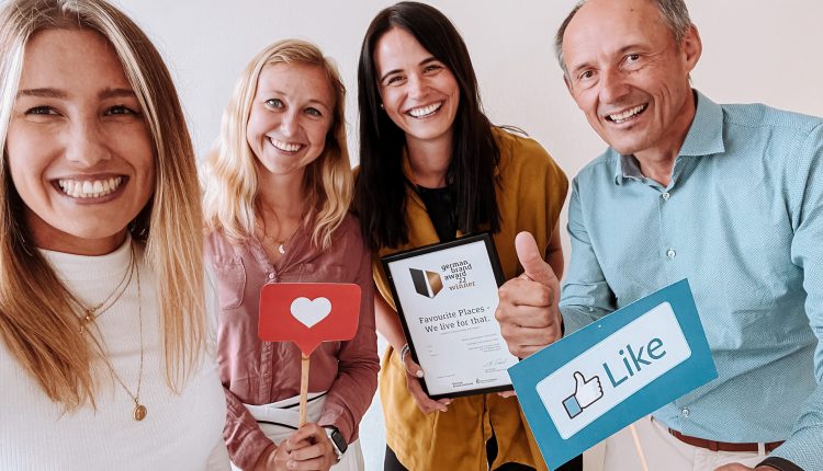 Vier lächelnde Menschen posieren mit einer Urkunde und Requisiten aus den sozialen Medien, darunter ein Herz und ein Like-Zeichen.