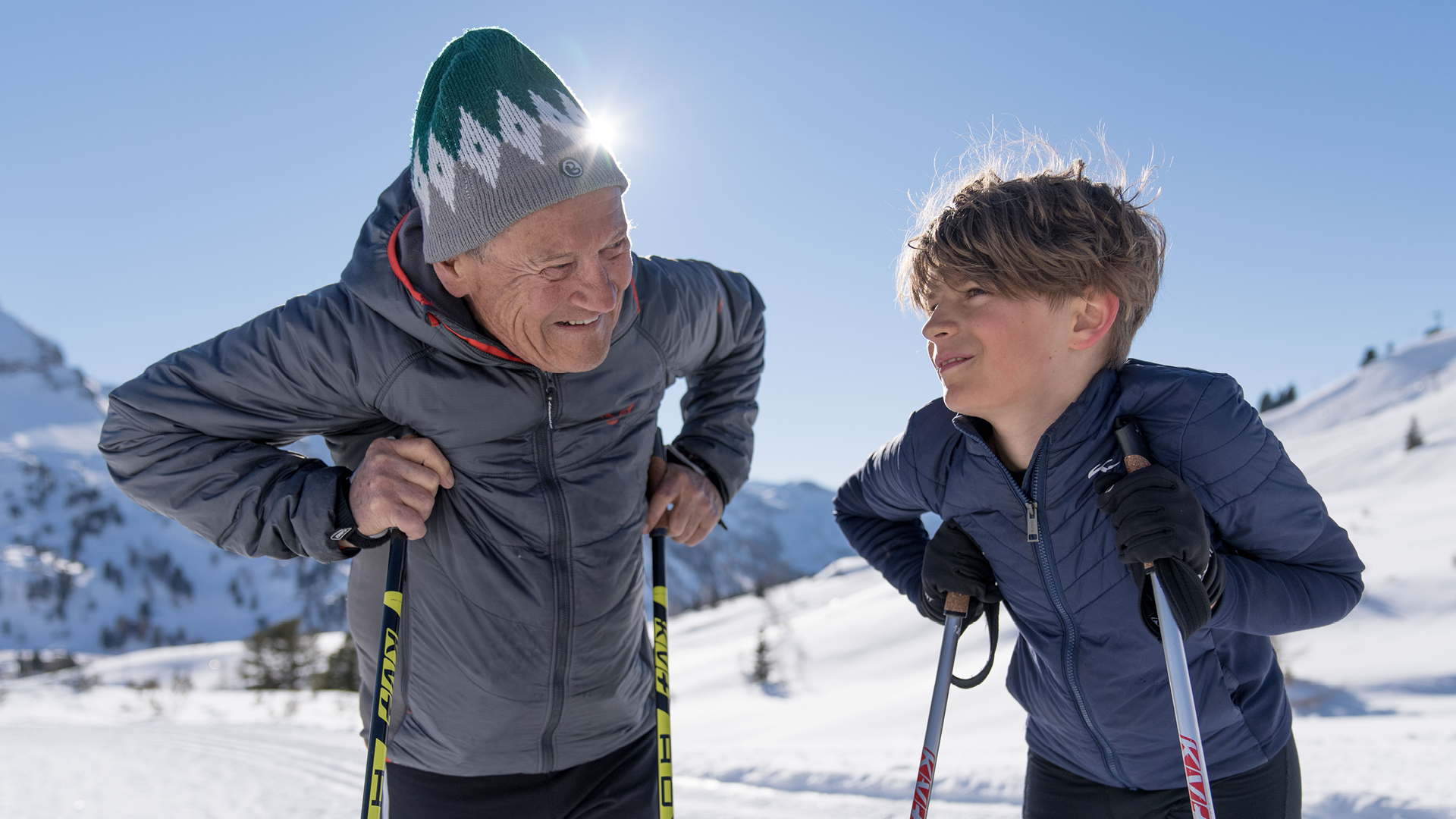 Ein älterer Mann und ein Junge lächeln sich beim Skilanglauf auf einem sonnigen, verschneiten Berg an.
