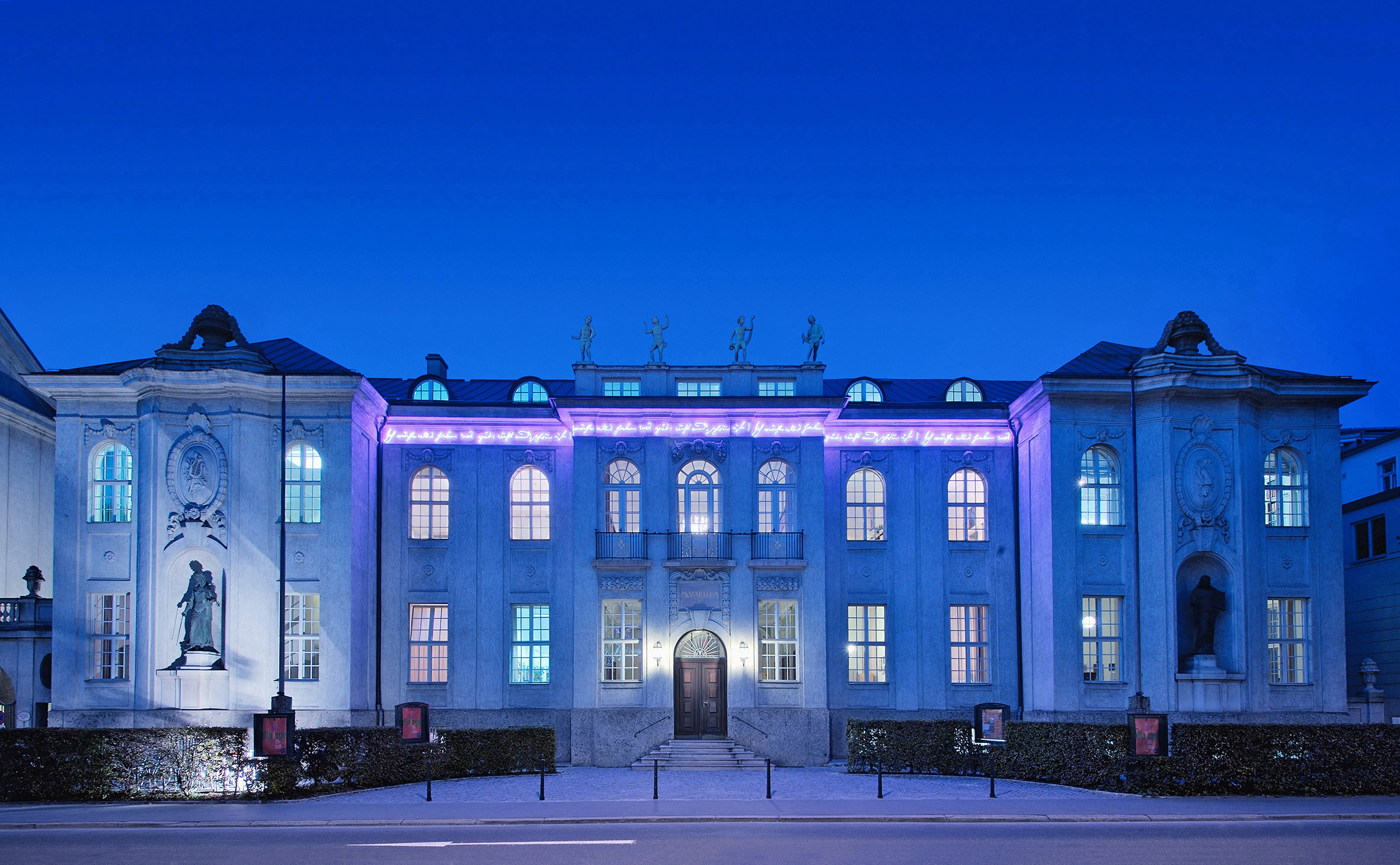 Elegantes Gebäude mit violetter Beleuchtung und Statuen, fotografiert in der Abenddämmerung vor einem tiefblauen Himmel.