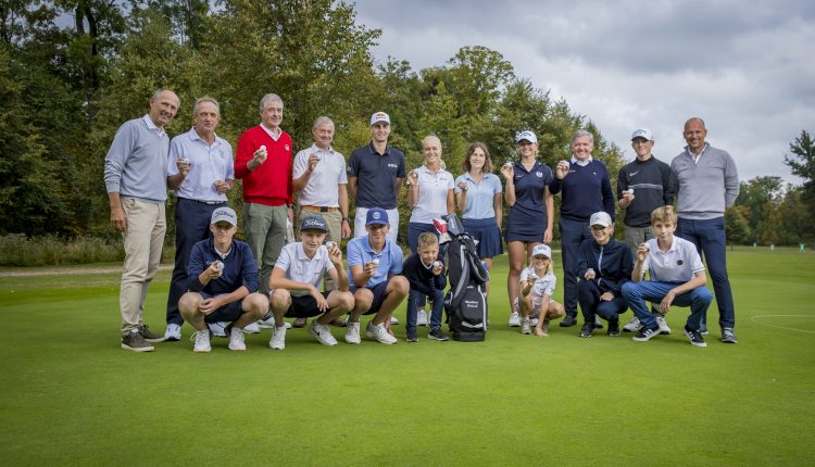 Eine Gruppe von Personen, darunter Kinder und Erwachsene, posieren gemeinsam auf einem Golfplatz und halten Trophäen.