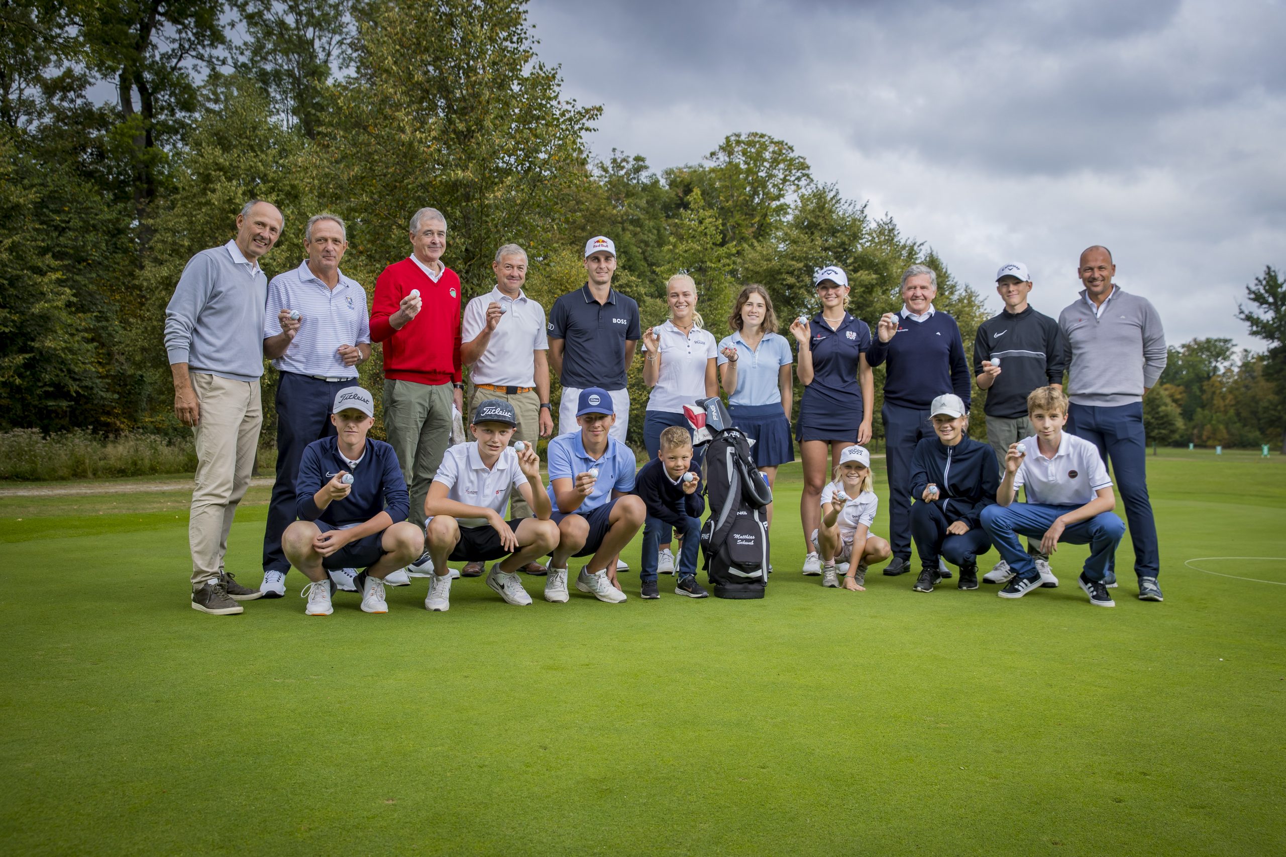 Eine Gruppe von Personen, darunter Kinder und Erwachsene, posieren gemeinsam auf einem Golfplatz und halten Trophäen.