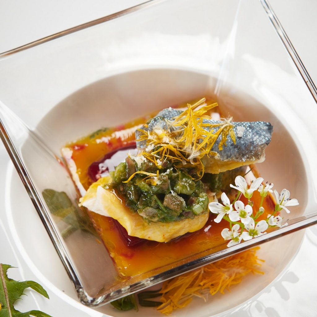 Ein Gourmet-Fischgericht mit Sauce, Grünzeug, essbaren Blüten und Garnitur in einer quadratischen Glasschale.