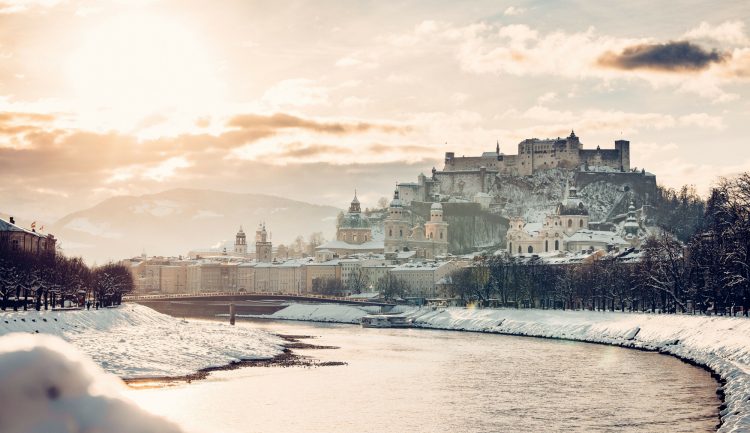 Verschneites Stadtbild von Salzburg mit einem Fluss, Kuppelkirchen und einer Festung auf dem Hügel bei Sonnenuntergang.