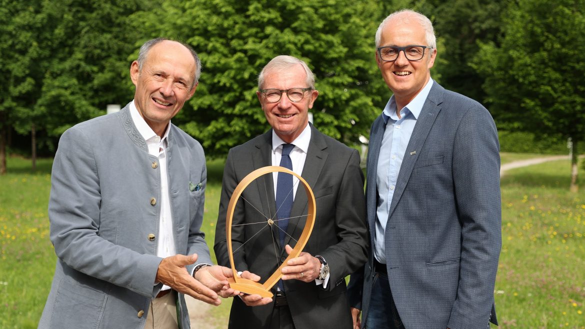 Drei lächelnde Männer in Anzügen stehen im Freien und halten einen hölzernen Preis mit einem radähnlichen Mittelstück.