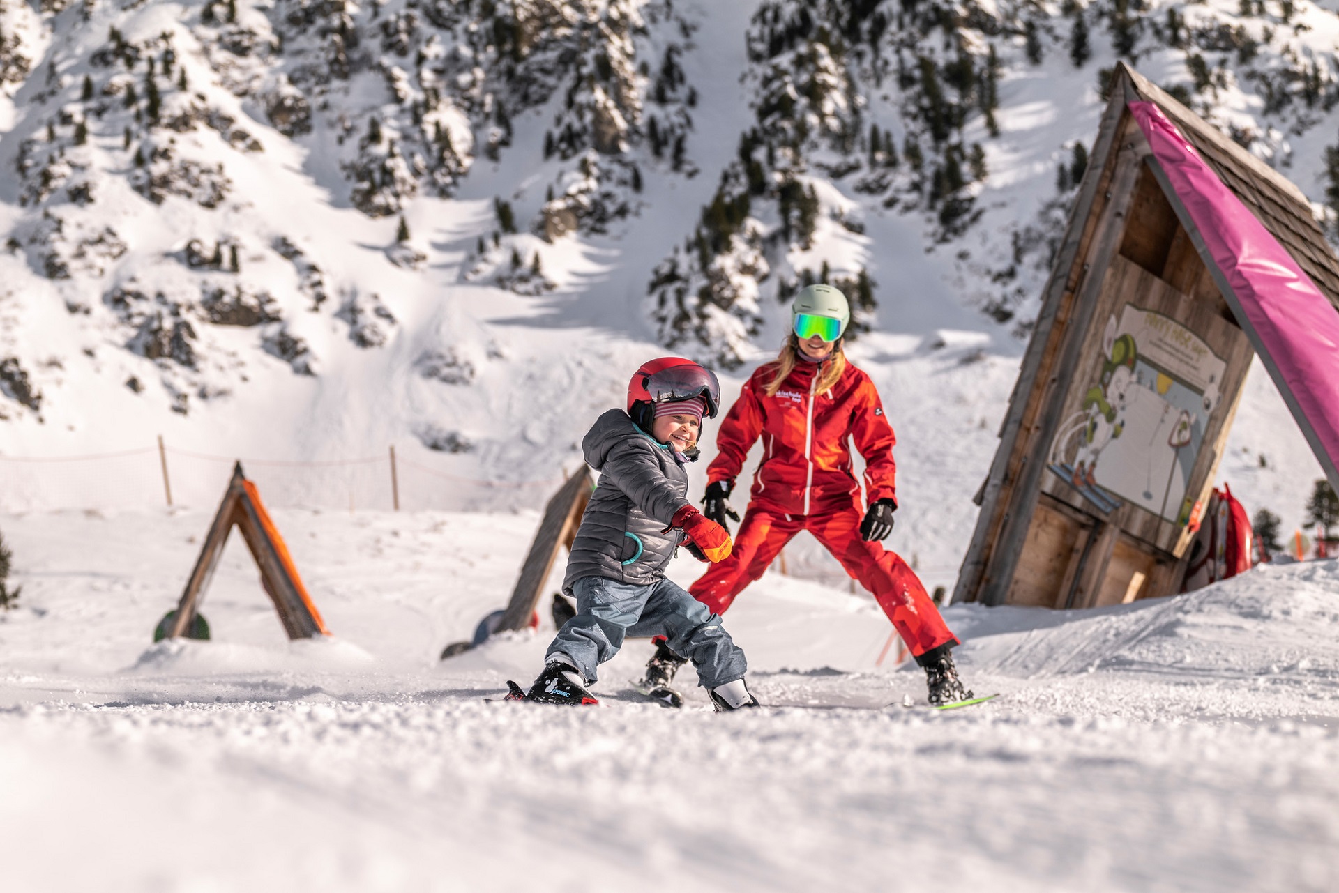 Ein Kind fährt auf Skiern einen verschneiten Hang hinunter, in der Nähe ein Skilehrer in Rot, beide tragen Helme und Skiausrüstung.