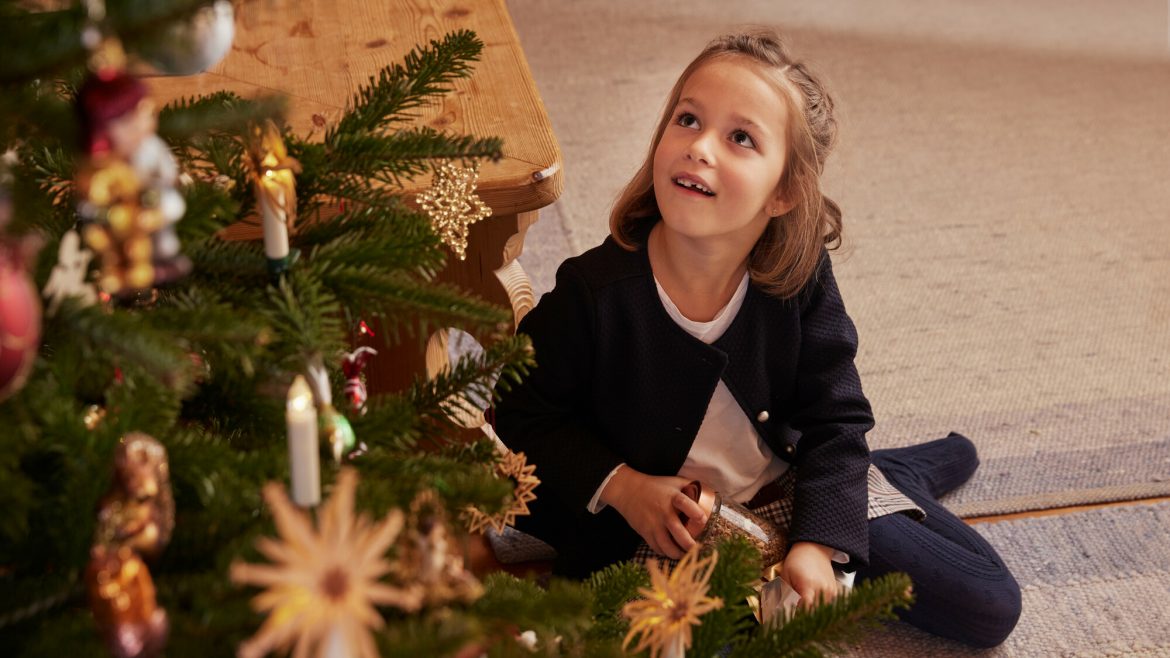 Ein junges Mädchen sitzt an einem geschmückten Weihnachtsbaum, lächelt und schaut aufgeregt nach oben.