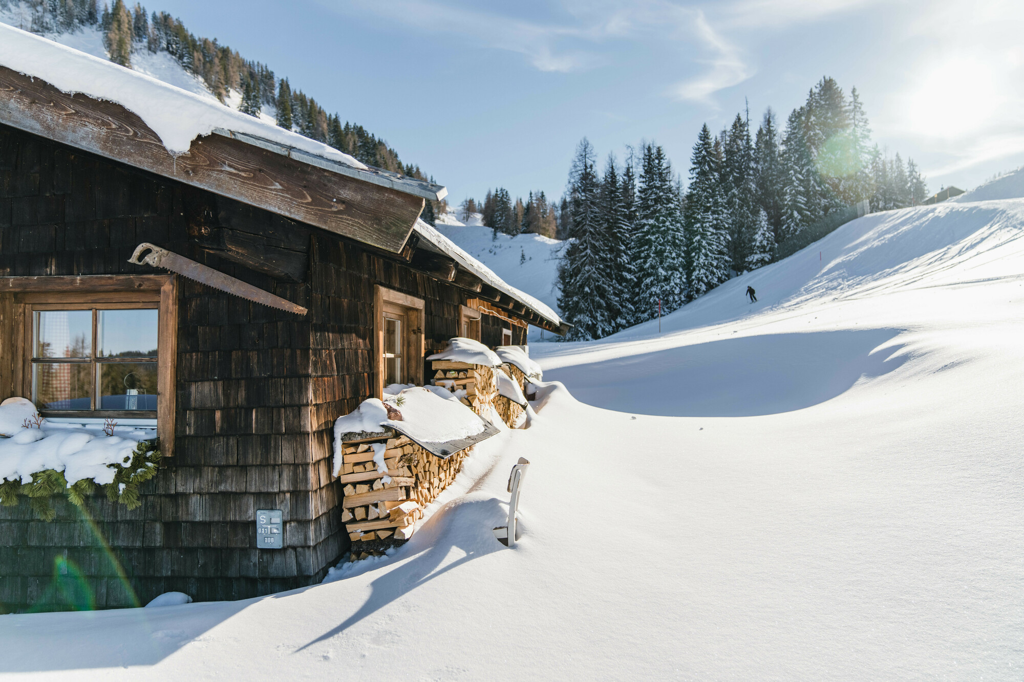 Eine Holzhütte inmitten von Tiefschnee und Kiefern, mit Sonnenlicht und einer Person, die in der Ferne Ski fährt.