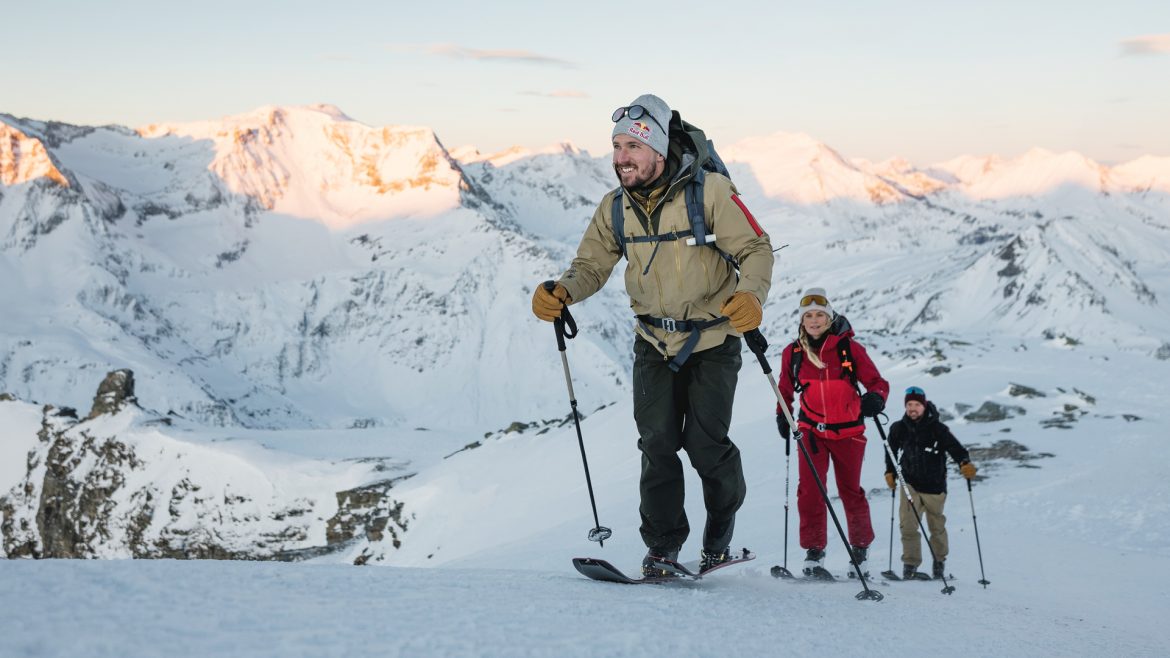Drei Personen in Winterausrüstung wandern mit Skistöcken einen verschneiten Berghang hinauf, umgeben von schneebedeckten Gipfeln.