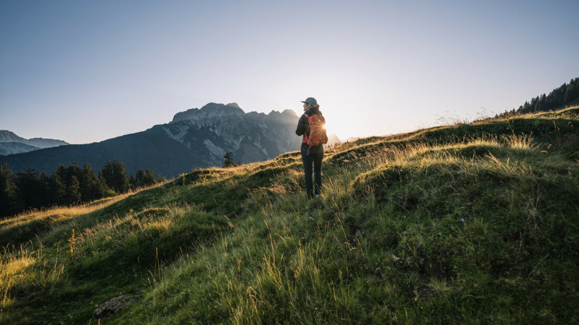 Ein Wanderer steht bei Sonnenaufgang auf einem grasbewachsenen Hügel, mit den Bergen im Hintergrund.