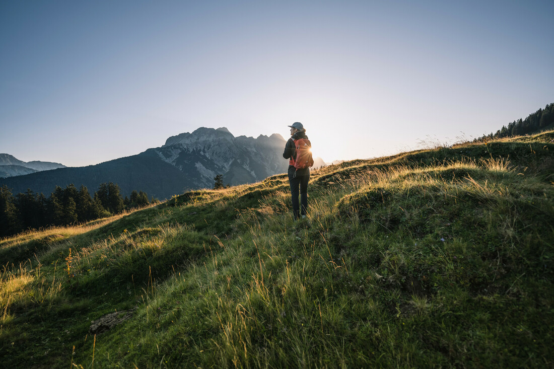 Ein Wanderer steht bei Sonnenaufgang auf einem grasbewachsenen Hügel, mit den Bergen im Hintergrund.