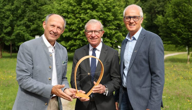 Drei Männer in Anzügen stehen im Freien, halten einen Holzpreis in der Hand und lächeln in die Kamera.