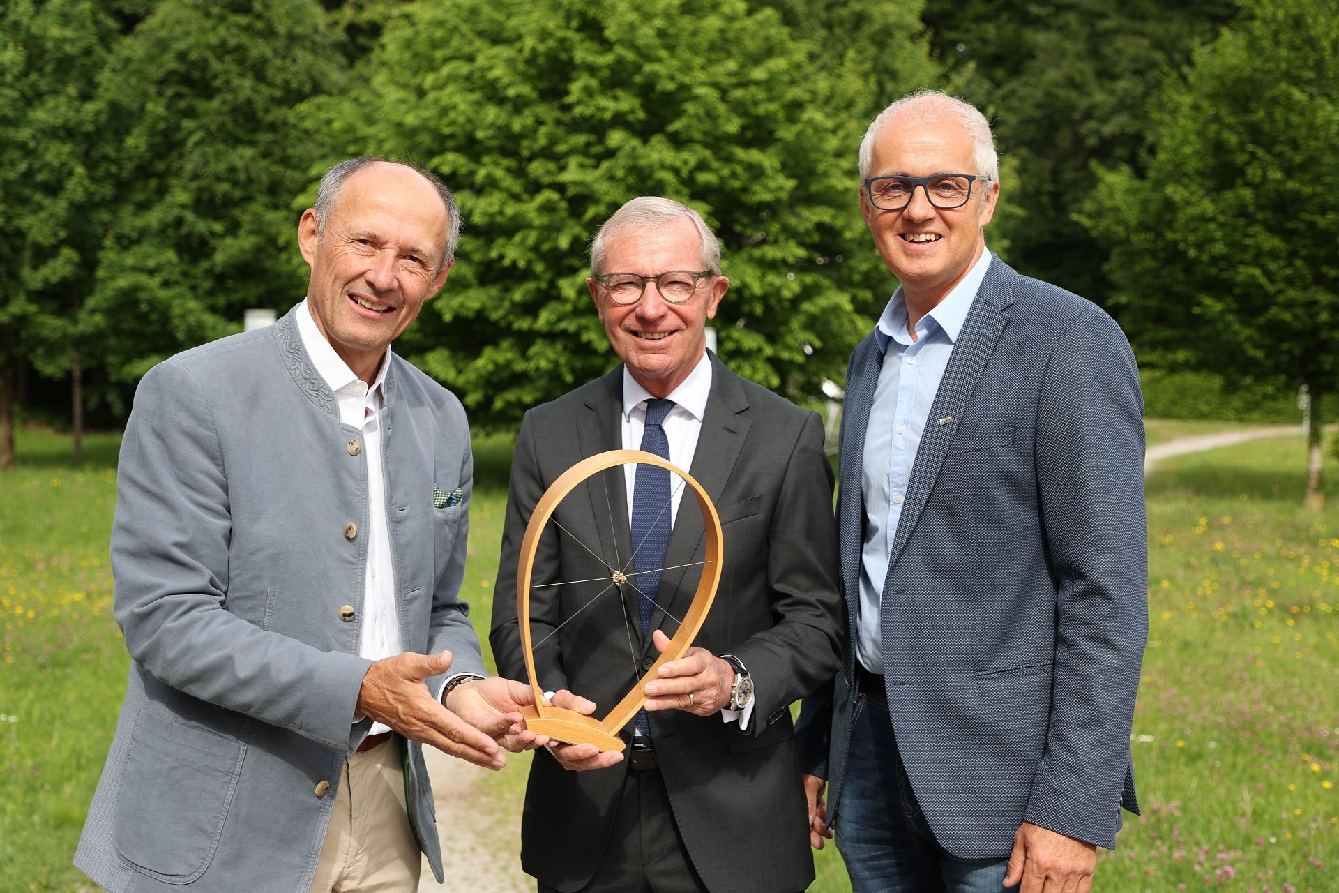 Drei Männer in Anzügen stehen im Freien, halten einen Holzpreis in der Hand und lächeln in die Kamera.