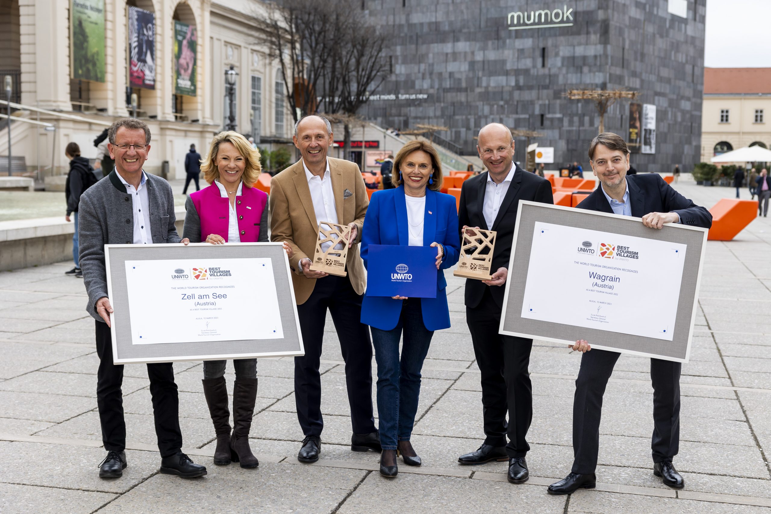 Sechs Personen stehen auf einem Stadtplatz mit Urkunden und Plaketten in der Hand und lächeln in die Kamera.