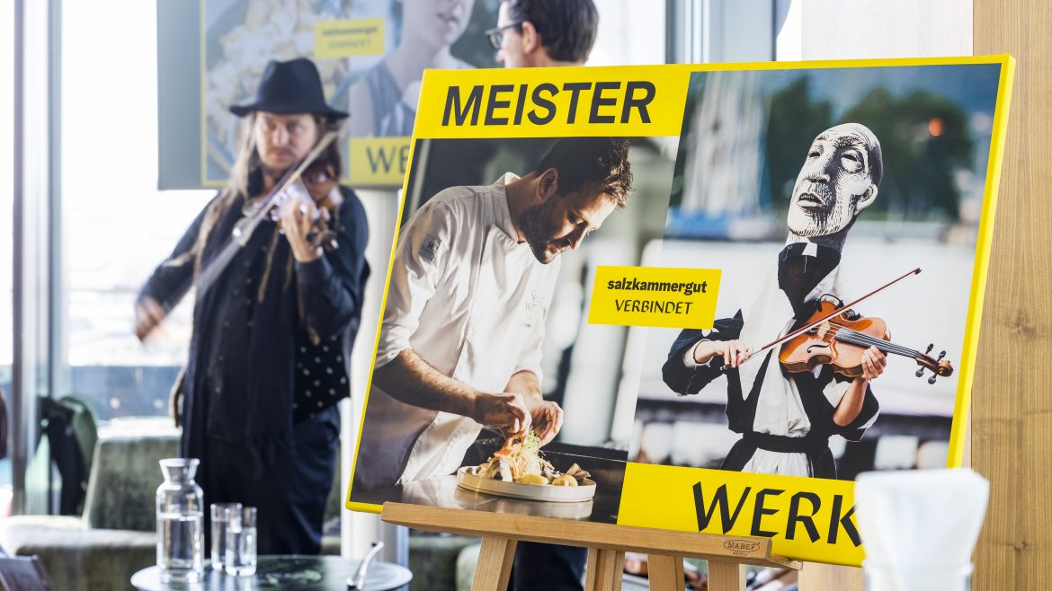 Ein Mann spielt Geige neben einem Plakat, das einen Koch und eine Geigenpuppe zeigt, mit dem Text MEISTER WERK.