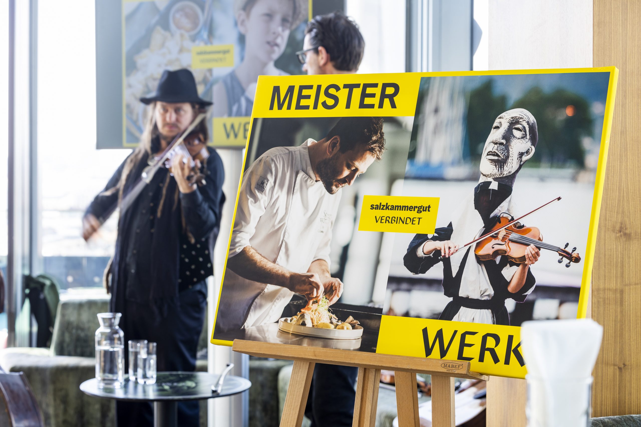 Ein Mann spielt Geige neben einem Plakat, das einen Koch und eine Geigenpuppe zeigt, mit dem Text MEISTER WERK.