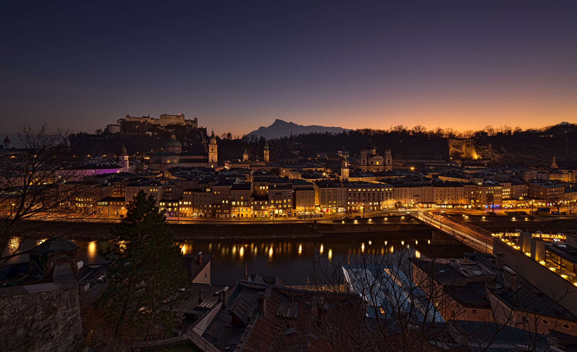 Eine Stadtlandschaft in der Abenddämmerung mit beleuchteten Gebäuden, einem Fluss und Bergen im Hintergrund.