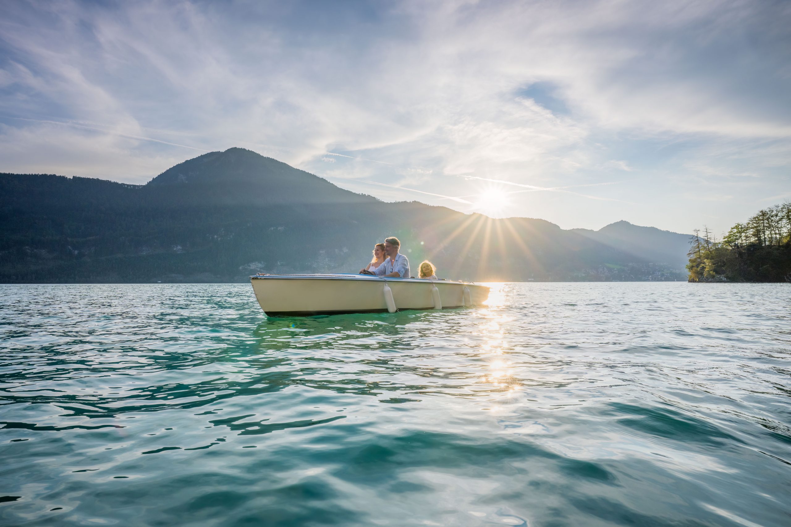 Eine dreiköpfige Familie in einem Ruderboot auf einem See mit Bergen und der untergehenden Sonne im Hintergrund.