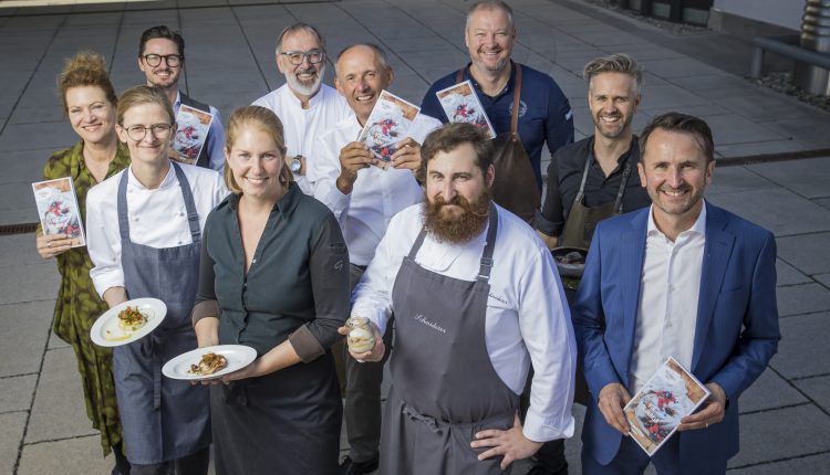 Eine Gruppe von lächelnden Köchen und Fachleuten posiert im Freien, einige halten Gerichte, andere Broschüren in der Hand.