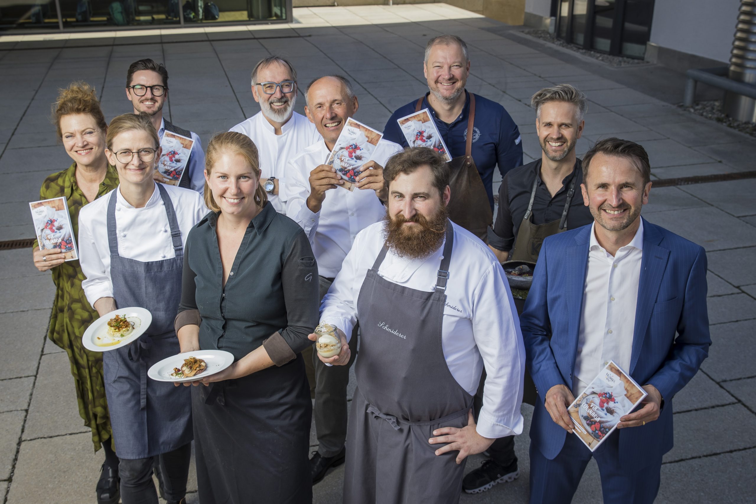 Eine Gruppe von lächelnden Köchen und Fachleuten posiert im Freien, einige halten Gerichte, andere Broschüren in der Hand.