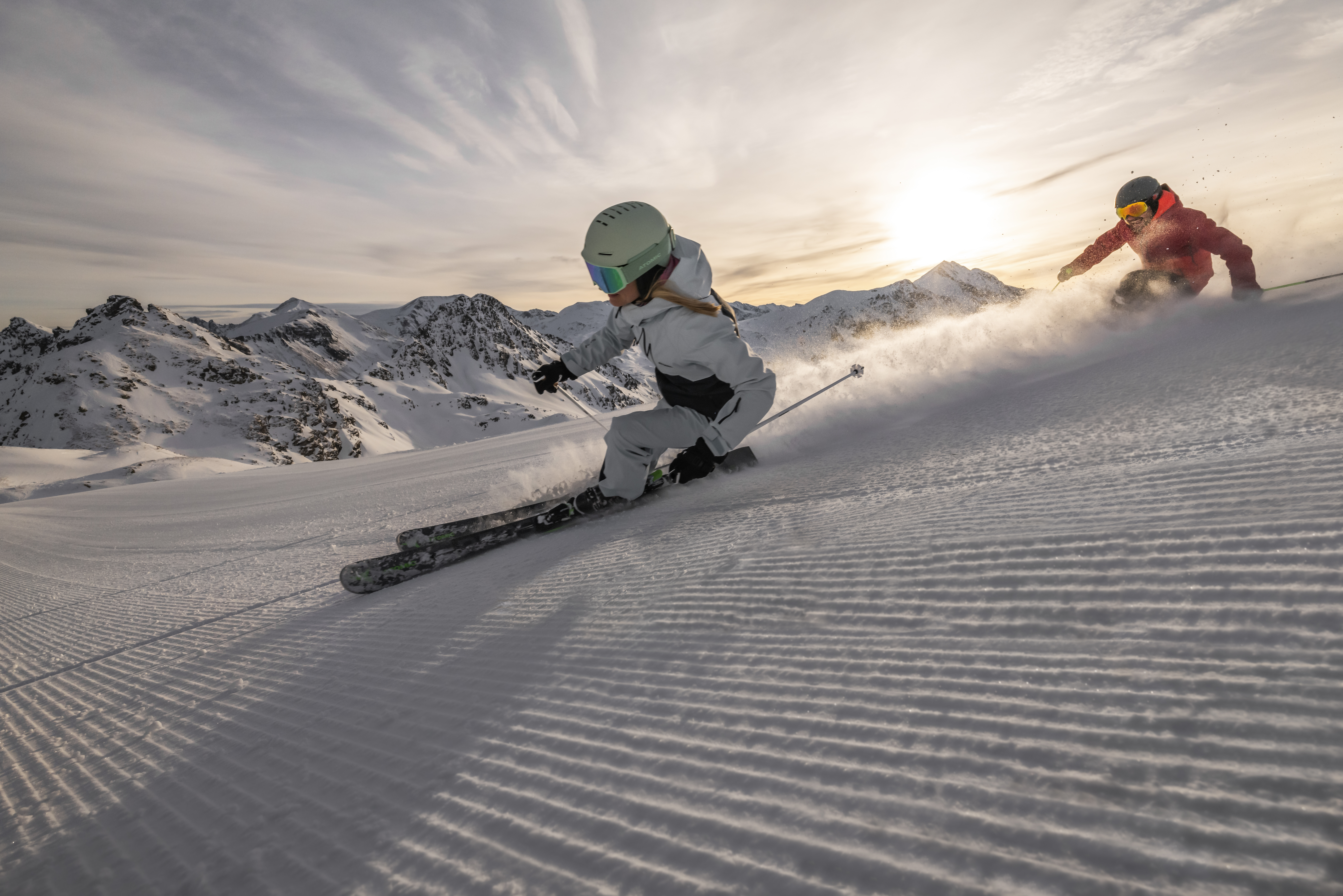 Zwei Skifahrer fahren auf einem verschneiten Berg mit Sonne und schroffen Gipfeln im Hintergrund bergab.