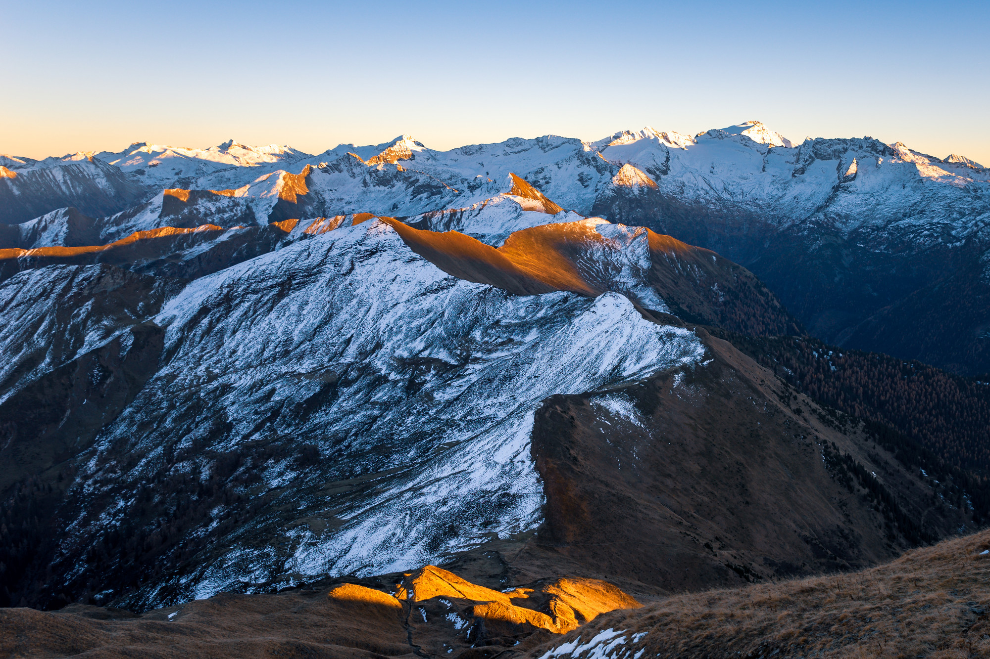Verschneite Berggipfel bei Sonnenaufgang mit goldenem Licht, das die Bergkämme und fernen Berge im Hintergrund hervorhebt.