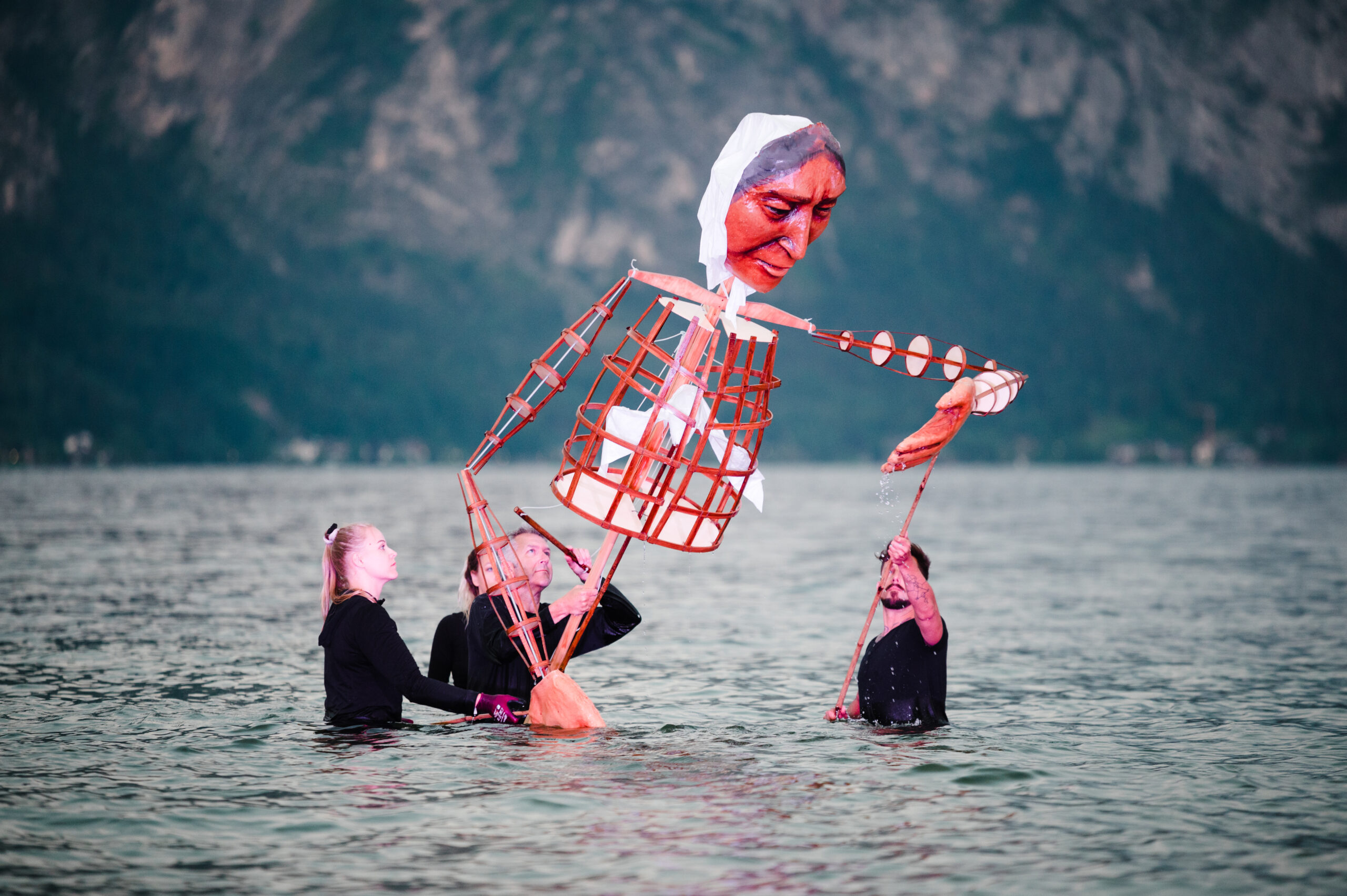 Drei Personen im Wasser bedienen eine große Puppe mit bemaltem Gesicht und Holzrahmen, im Hintergrund Berge.