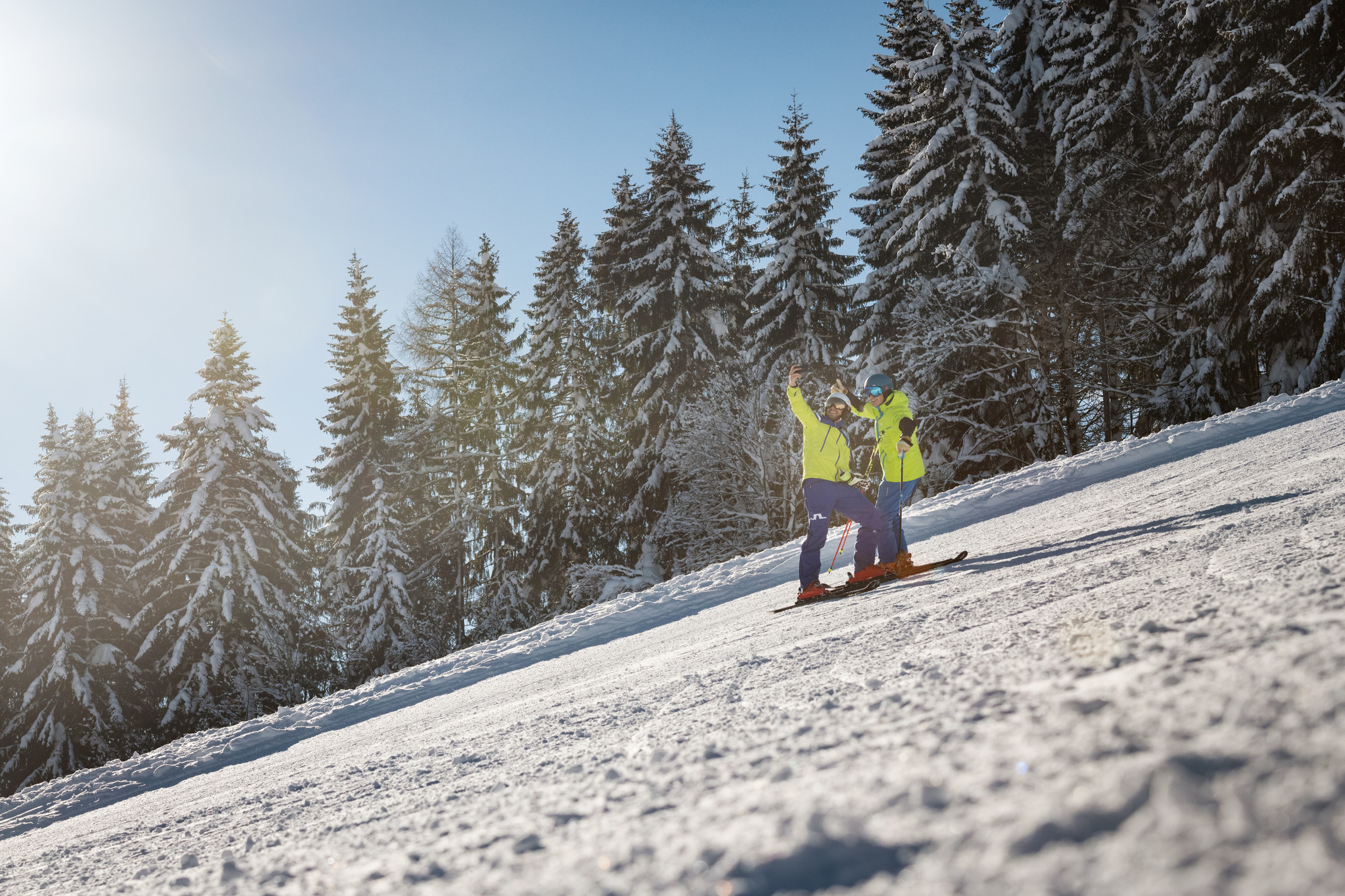 Zwei Personen in heller Skiausrüstung stehen auf einer verschneiten Piste mit Bäumen und Sonnenlicht im Hintergrund.