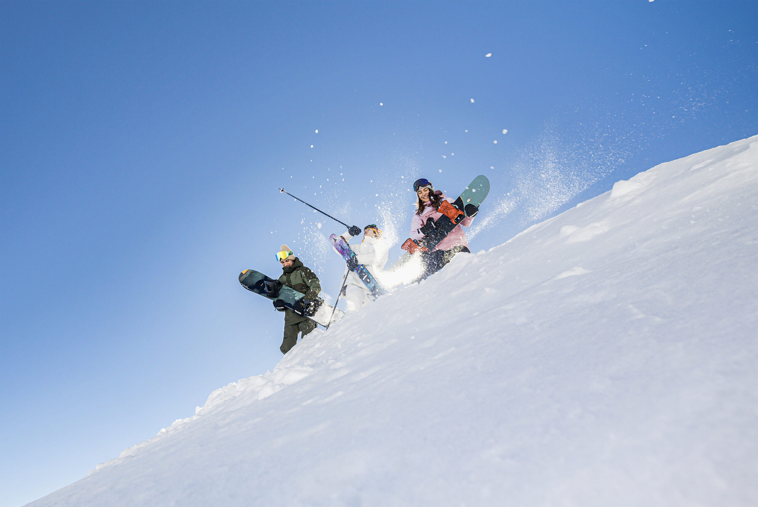 Drei Personen mit Snowboards gehen einen verschneiten Hang hinauf, unter einem klaren blauen Himmel, das Sonnenlicht scheint hinter ihnen.