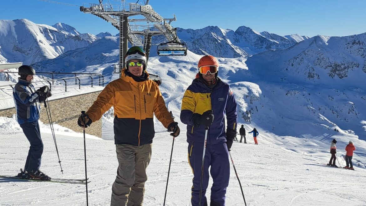 Zwei Skifahrer posieren an einem sonnigen Tag auf einer verschneiten Piste mit Bergen und einem Skilift im Hintergrund.