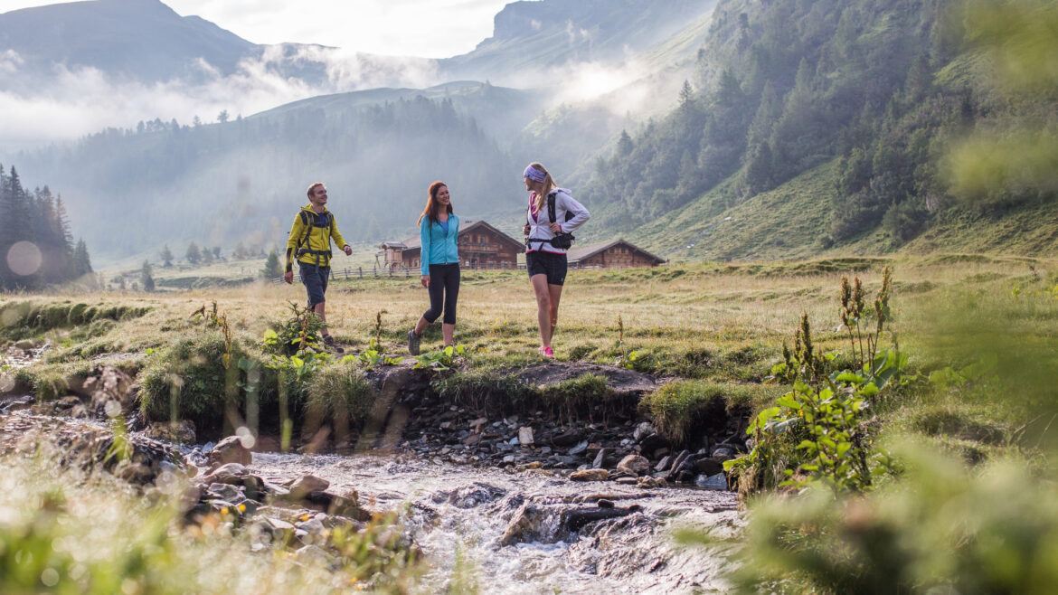 Drei Personen wandern entlang eines Baches in einer grasbewachsenen Berglandschaft mit Nebel und Hütten im Hintergrund.