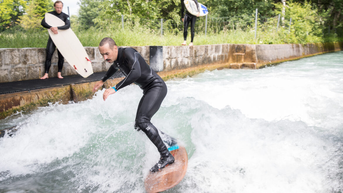Ein Mann surft auf einer Flusswelle, während zwei Personen mit Surfbrettern vom Betonufer aus zusehen.