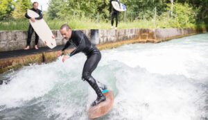 Ein Mann surft auf einer Flusswelle, während zwei Personen mit Surfbrettern vom Betonufer aus zusehen.